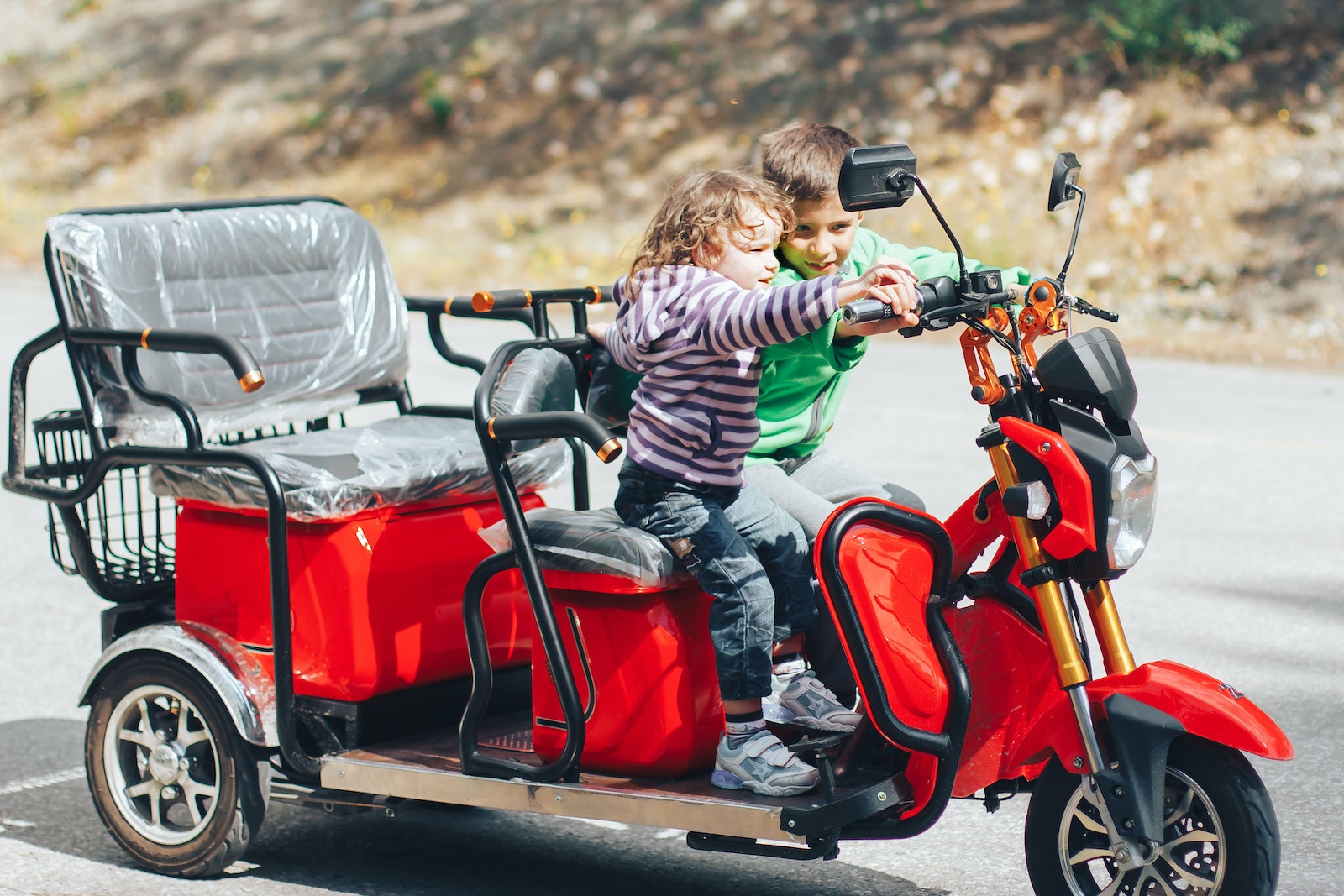 Adorable child helping little brother to ride modern red trike motorcycle on asphalt road on sunny day